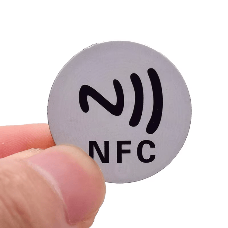 لقطة مقربة لوسم RFID دائري رمادي من PVC مخصص للأسطح المعدنية ويحمل علامة NFC، ممسوك بين الأصابع لإظهار الشكل المدمج للملصق العازل لتأثير المعدن.